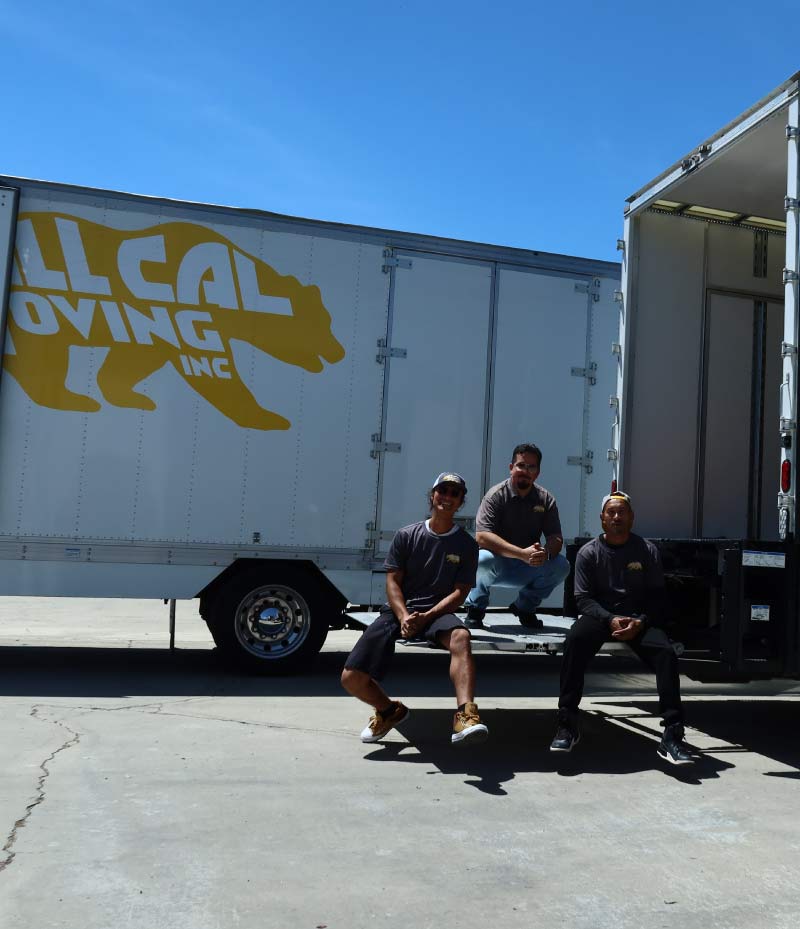 Three professional All Cal Moving crew members posing on the lift gate of a moving truck with the gold bear logo on a sunny day in Visalia, ready for a residential move